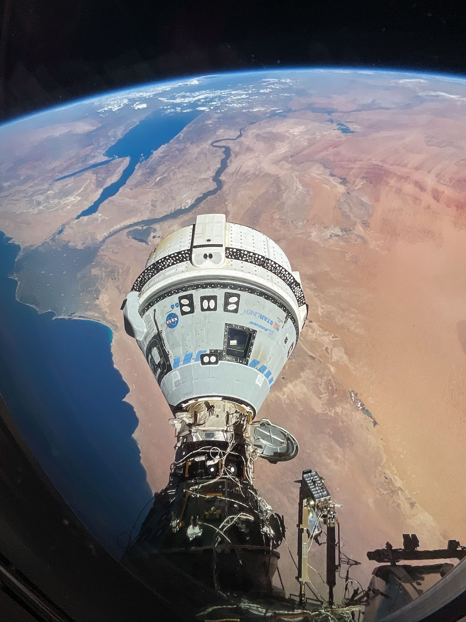 nave espacial boeing starliner em forma de lágrima acoplada a um grande tubo coberto de arame na lateral da estação espacial acima da terra com um continente arenoso marrom e vermelho se estendendo abaixo