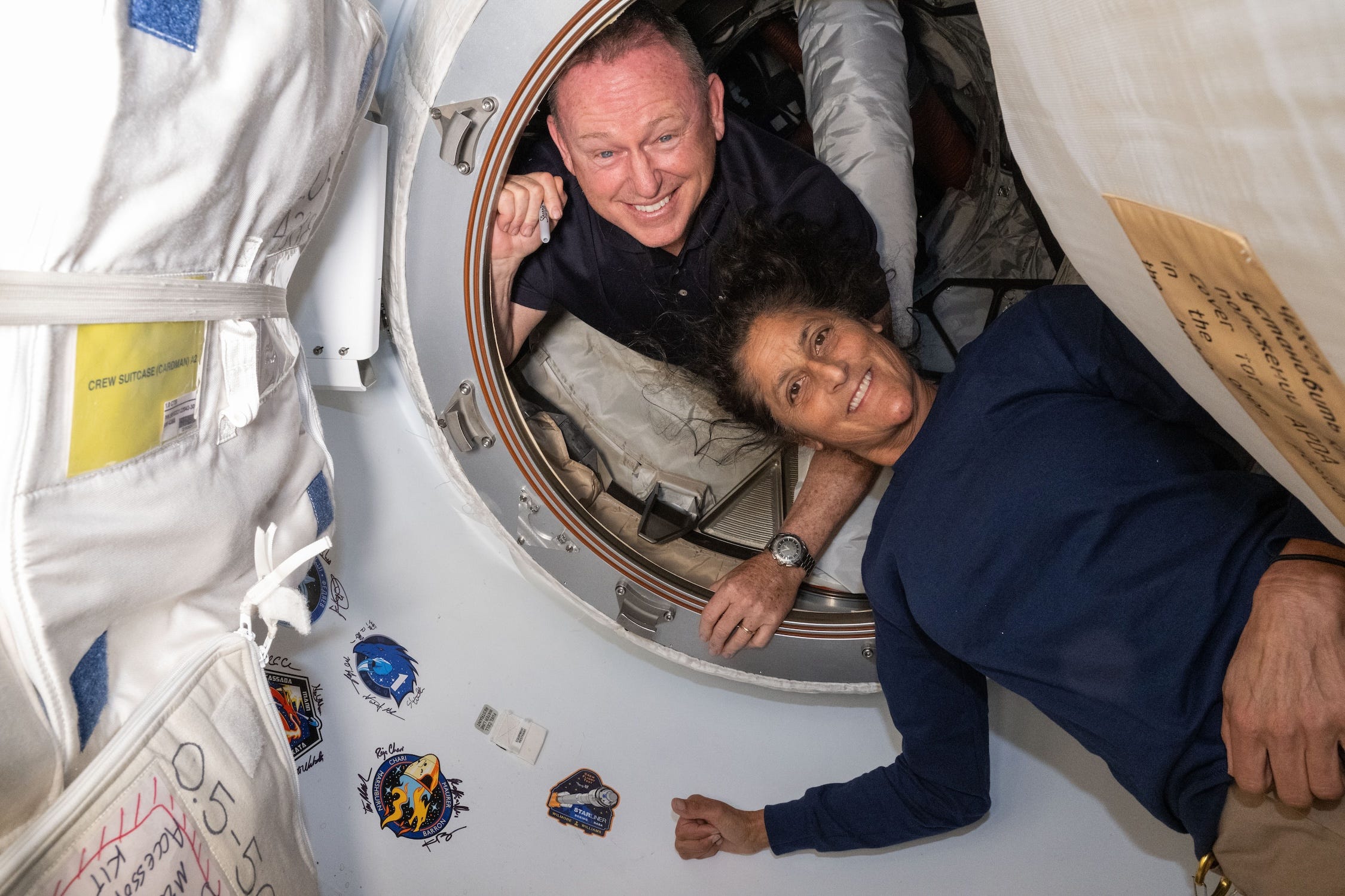 sorridentes Butch Wilmore e Suni Williams flutuando em um túnel circular branco ao redor de um porto na estação espacial