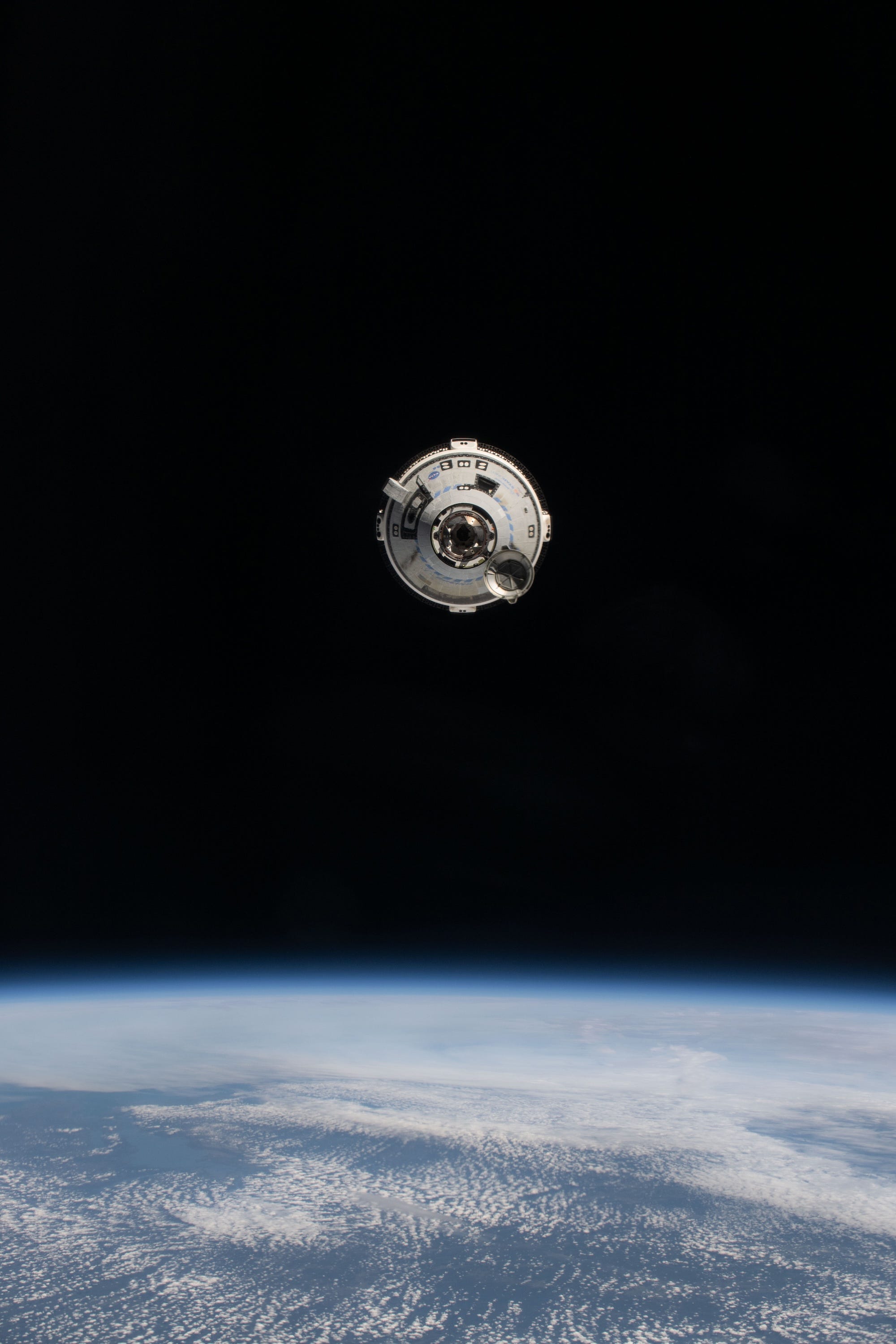 A nave espacial Boeing Starliner vista de frente com o nariz aberto e o portal de ancoragem visível na escuridão do espaço acima da curvatura azul e nublada da Terra