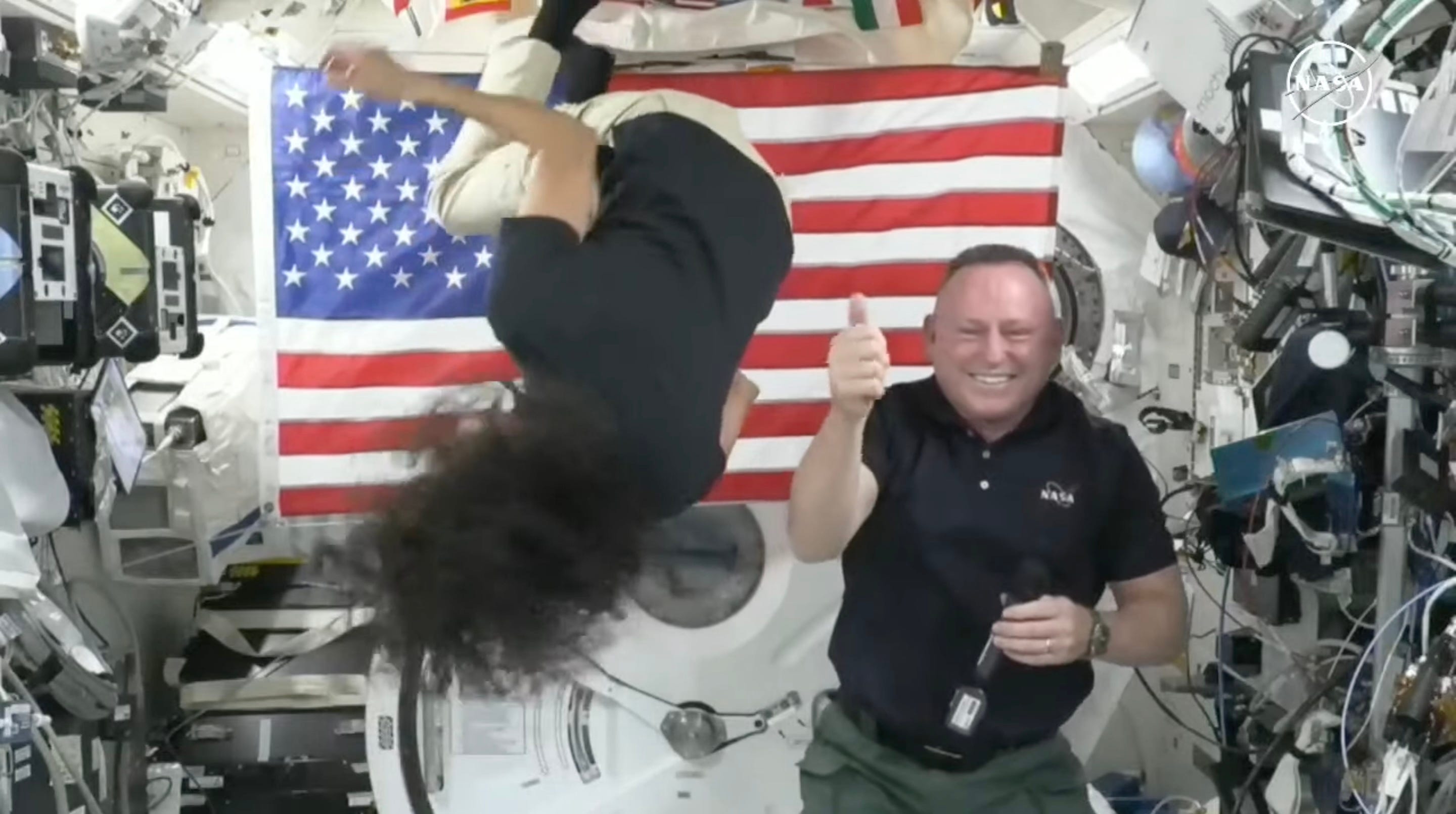 a astronauta suni williams de cabeça para baixo girando perto do teto da estação espacial enquanto Butch Wilmore levanta o polegar sorrindo ao lado dela na frente de uma bandeira americana ao lado de paredes cobertas de gadgets