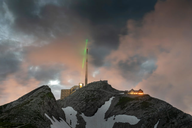 Laser verde irradiando para o céu nublado acima da torre de telecomunicações no cume de uma montanha coberta de neve.