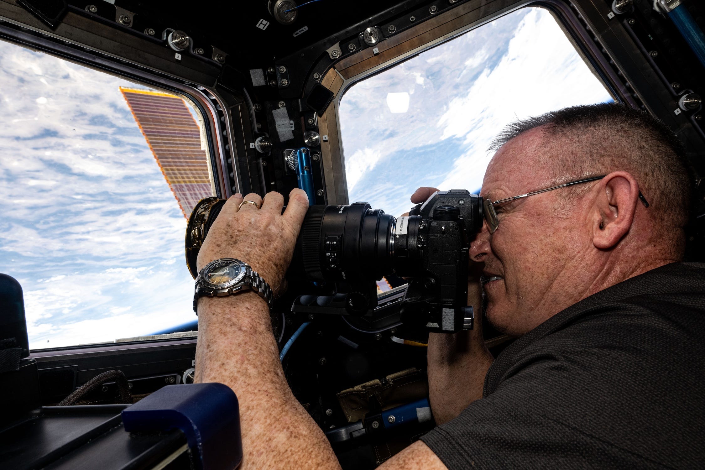 Butch Wilmore olhando através de uma câmera, ele está apontando para uma janela retangular dentro da estação espacial olhando para a terra azul e nublada abaixo