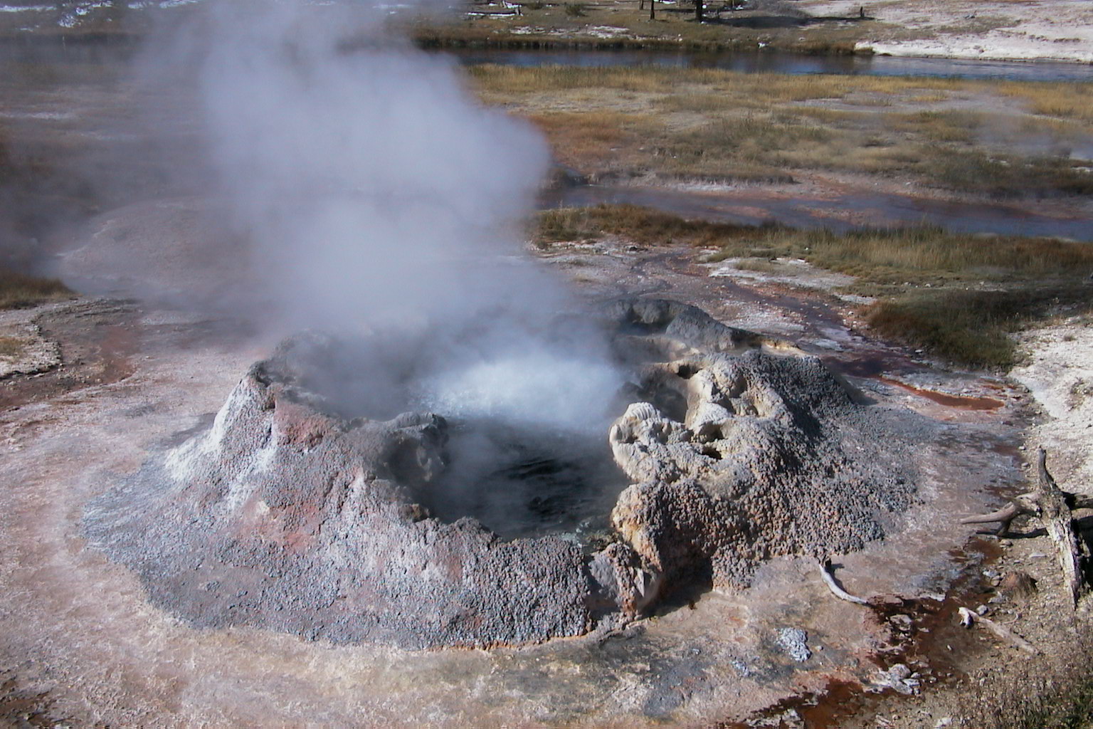 Uma fonte termal com paredes minerais cinza naturalmente formadas em torno de sua borda, cercada por um chão rochoso plano que parece levemente musgo. A fonte termal tem vapor de seu centro.